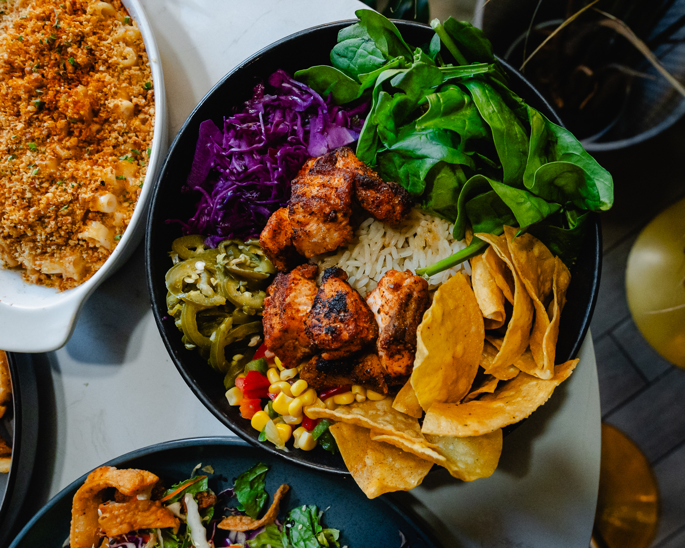 Blackened salmon grain bowl in a dark ceramic bowl with rice, greens, purple cabbage, corn, jalapeños and tortilla chips