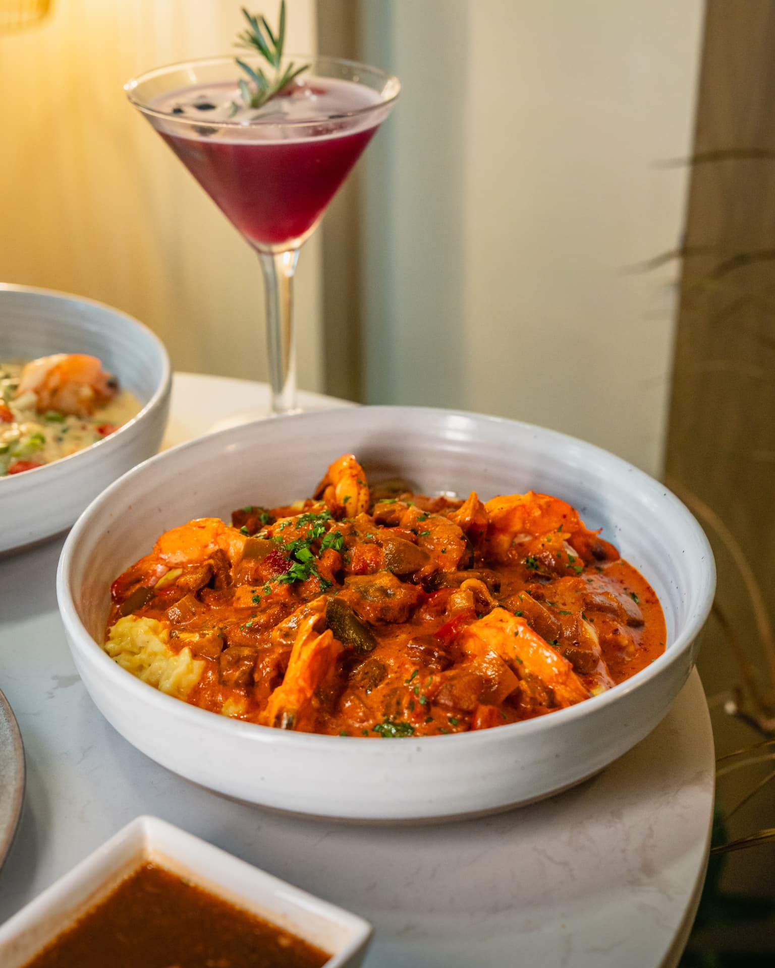 Stone-ground shrimp and grits in a wide ceramic bowl, garnished with herbs, with a cocktail behind